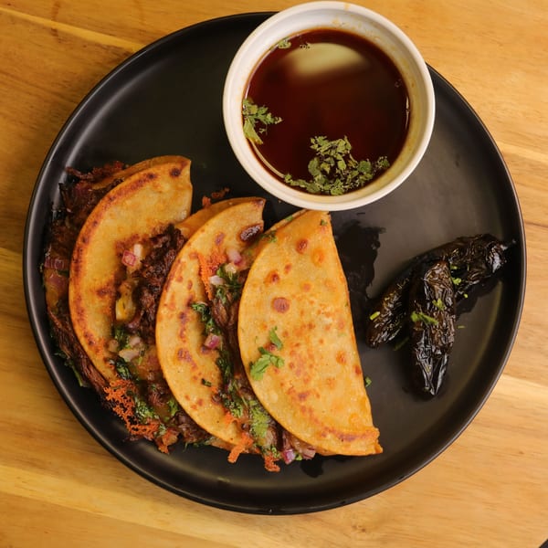 Three birria tacos with cilantro and onion, a side of consommé, and roasted peppers.