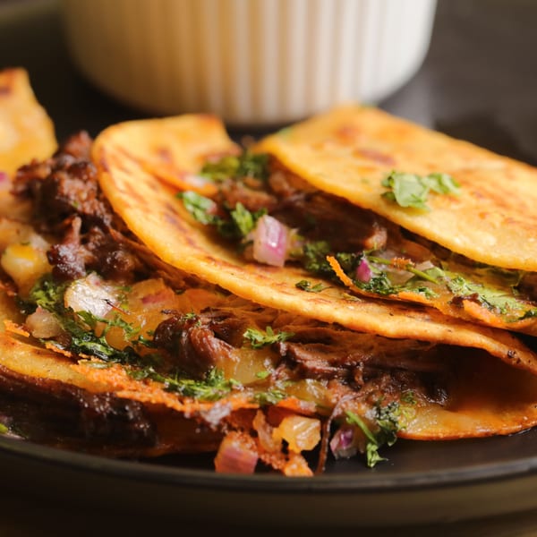 Close-up of three Bueno Burrito birria tacos with melted cheese, cilantro, and red onion.