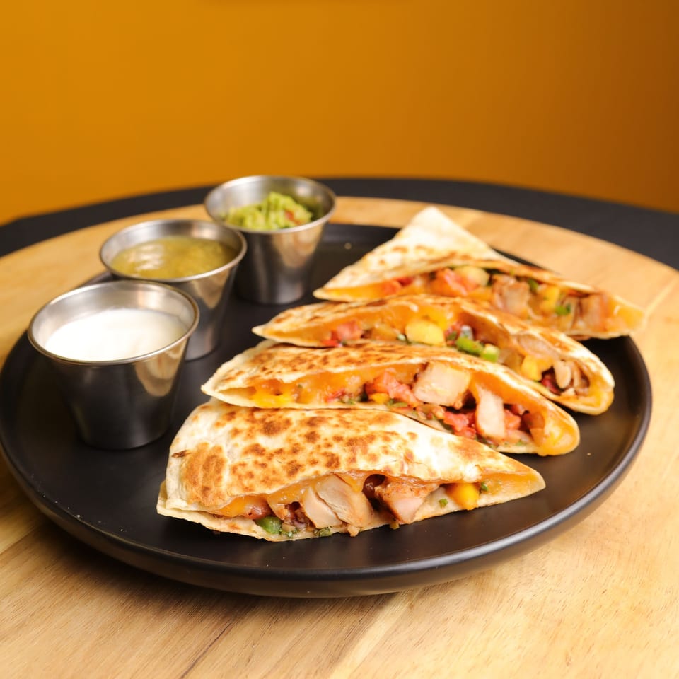 Four chicken quesadilla slices with melted cheese and salsa, served with sour cream, salsa verde, and guacamole.