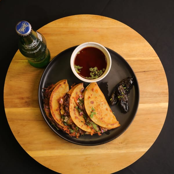 Three birria tacos with a side of consommé, grilled peppers, and a Sprite on a wooden board.