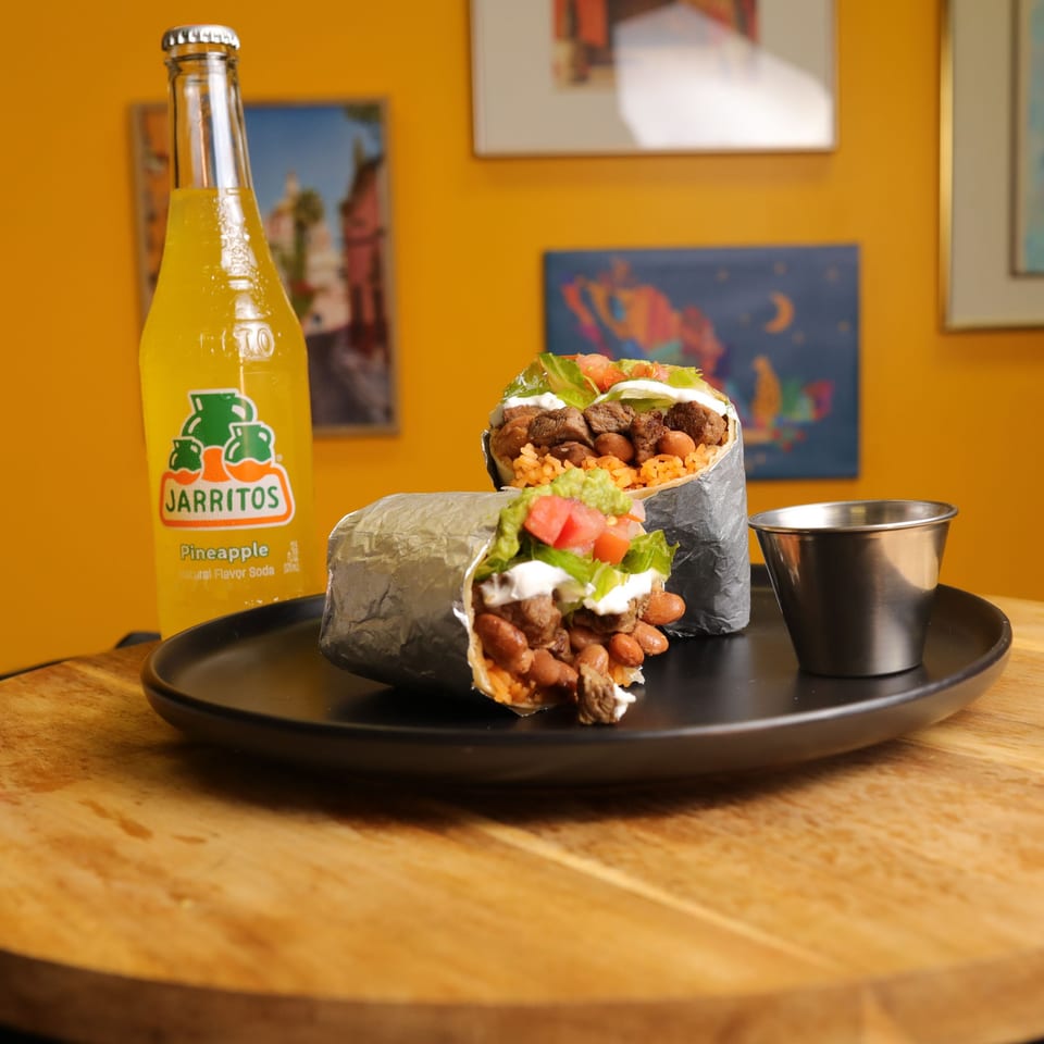 Two Bueno Burrito burritos with meat, beans, rice, and toppings, alongside a pineapple Jarritos soda.